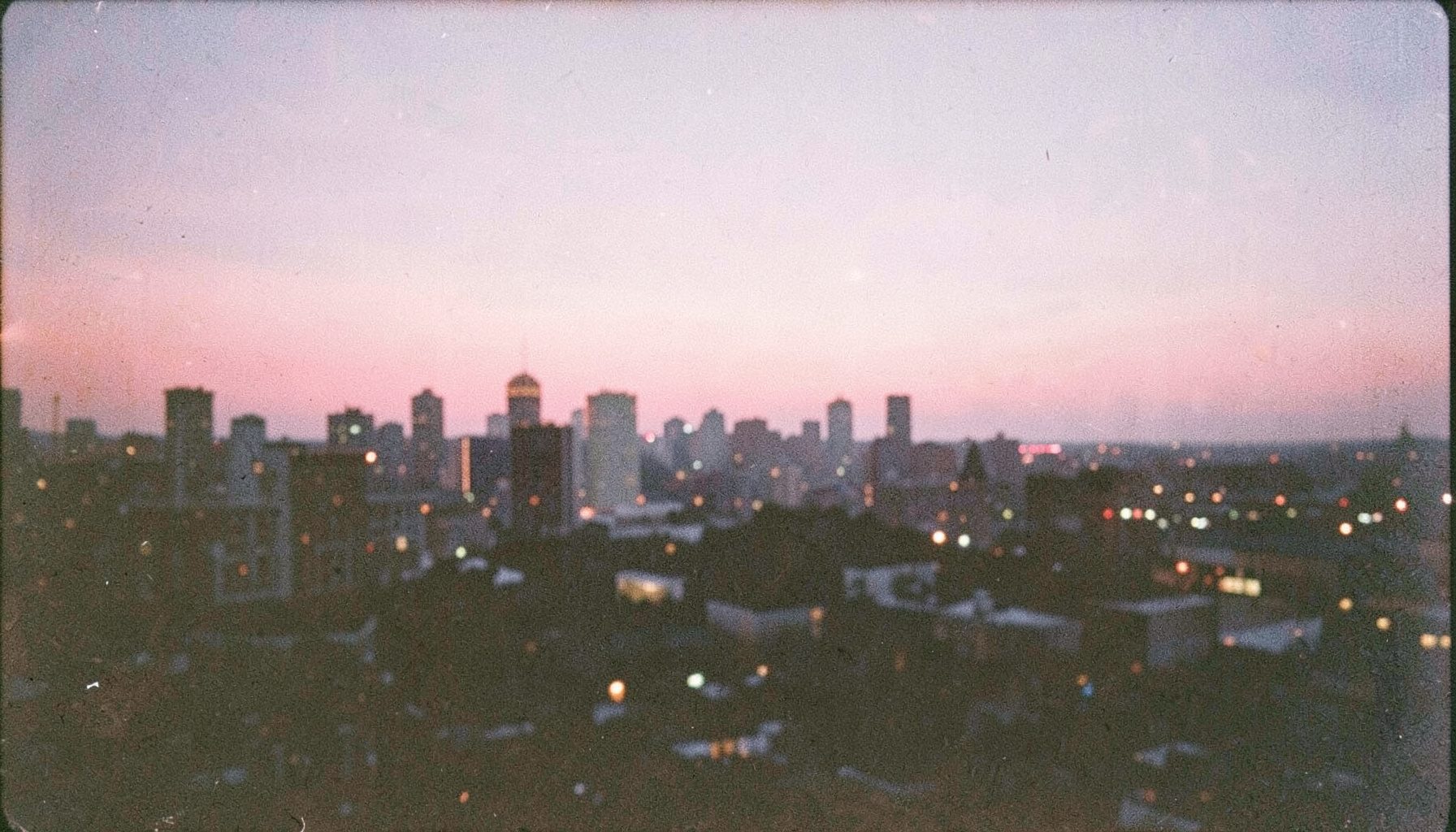 Analog film photograph of a softly lit city skyline at dusk, with faded pink highlights, warm grain and a quiet romantic mood