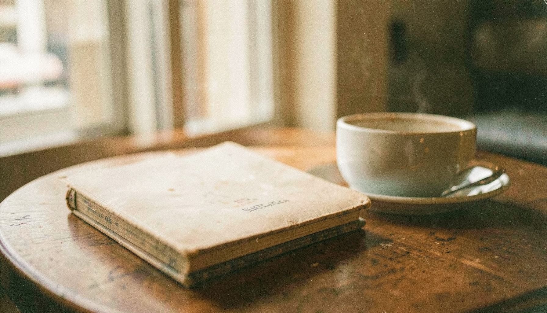 A worn paperback book resting on a cafe table beside an espresso cup, soft morning light through a window, muted browns