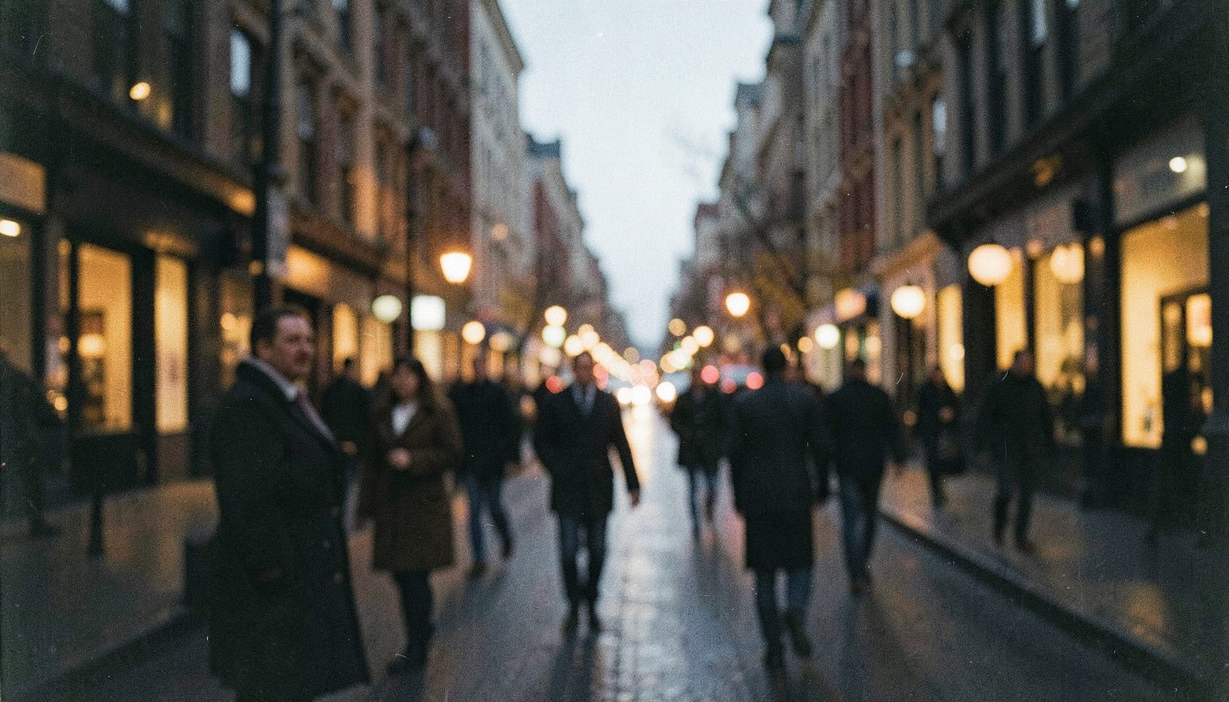 Softly lit city street at dusk with blurred pedestrians in coats, warm amber and deep navy tones, grainy film photograph